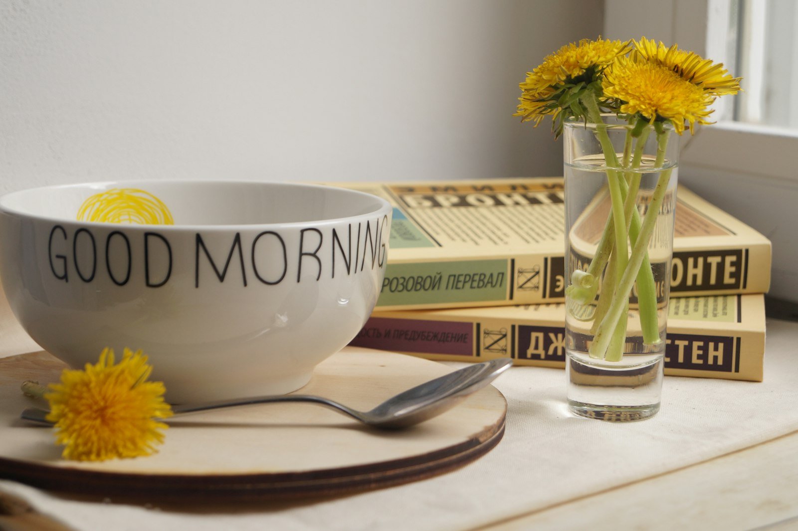 A white bowl with Good Morning text, dandelions in a glass, and books